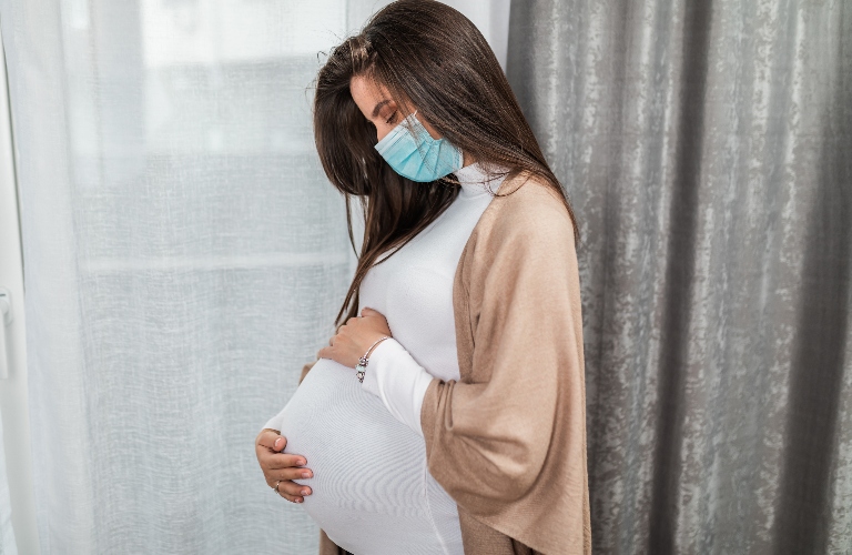 Young pregnant woman is standing by window with face protective mask