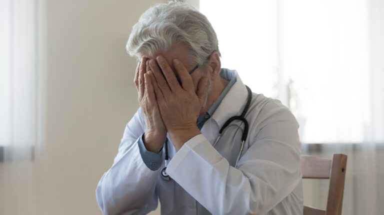 doctor wearing white uniform with stethoscope covering face with hands