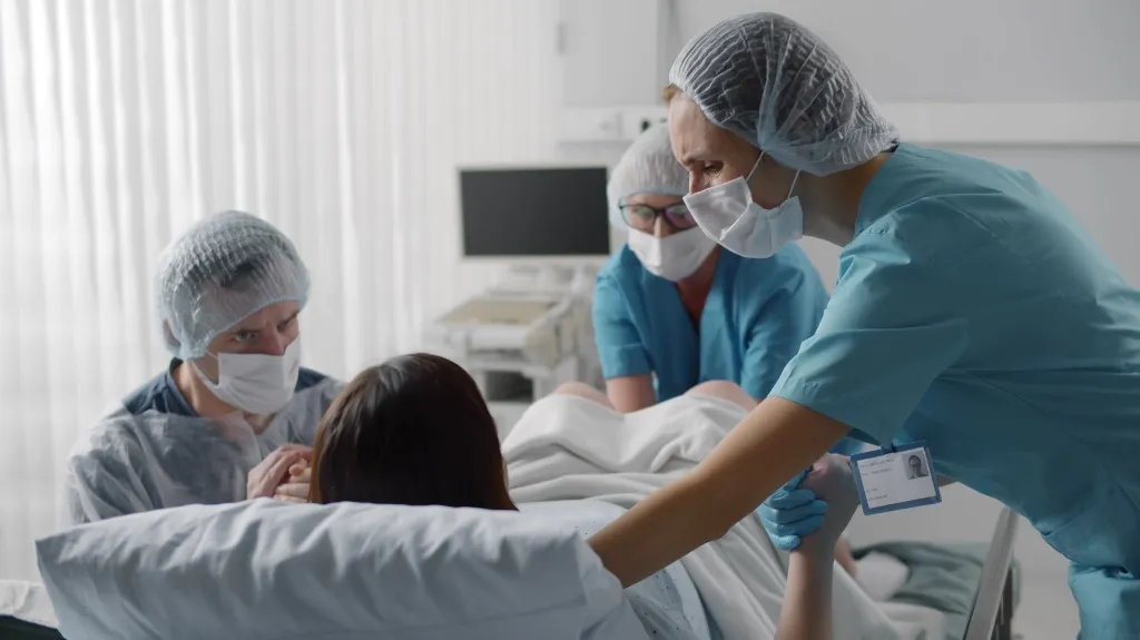 Medical professionals in blue scrubs and masks assisting a patient in a hospital bed.